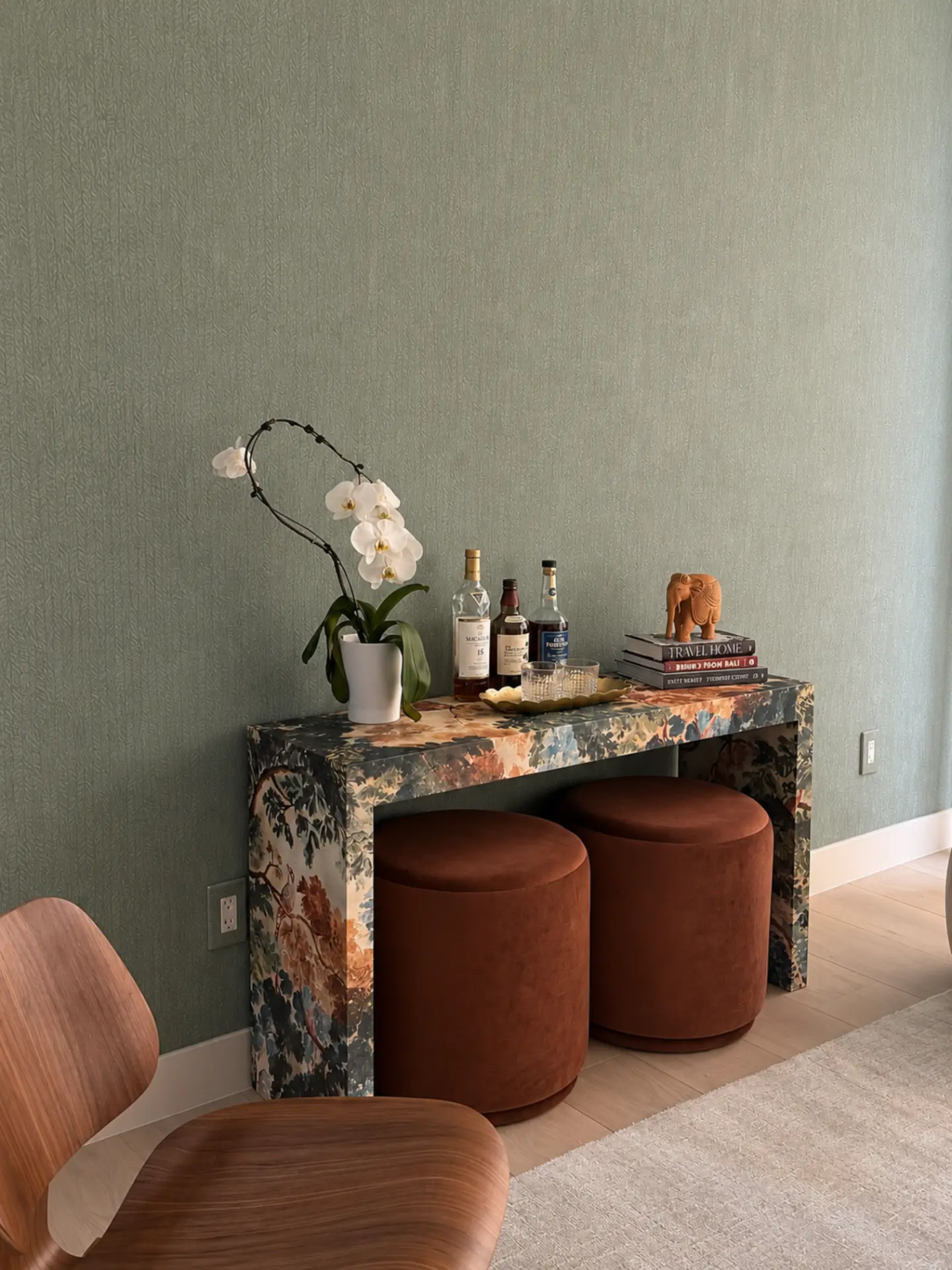 Styled console table wrapped in dark botanical fabric against sage patterned wallpaper, with two rust velvet stools tucked beneath, a white orchid in a ceramic pot, liquor bottles including a Macallan 15, a gold tray with glass tumblers, and a stack of books topped with a carved wooden elephant