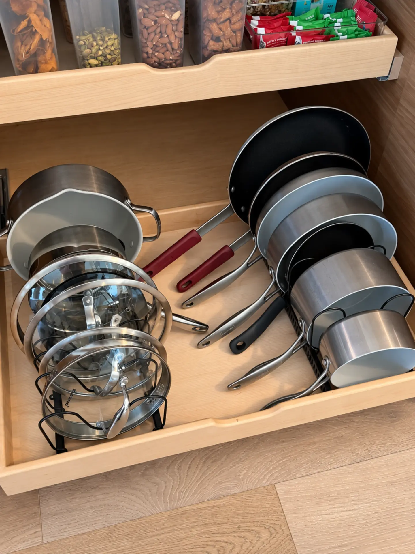 Pantry pull-out shelf with bins of snacks and nuts above a rack holding stainless steel pots, red-handled pans, and glass lids