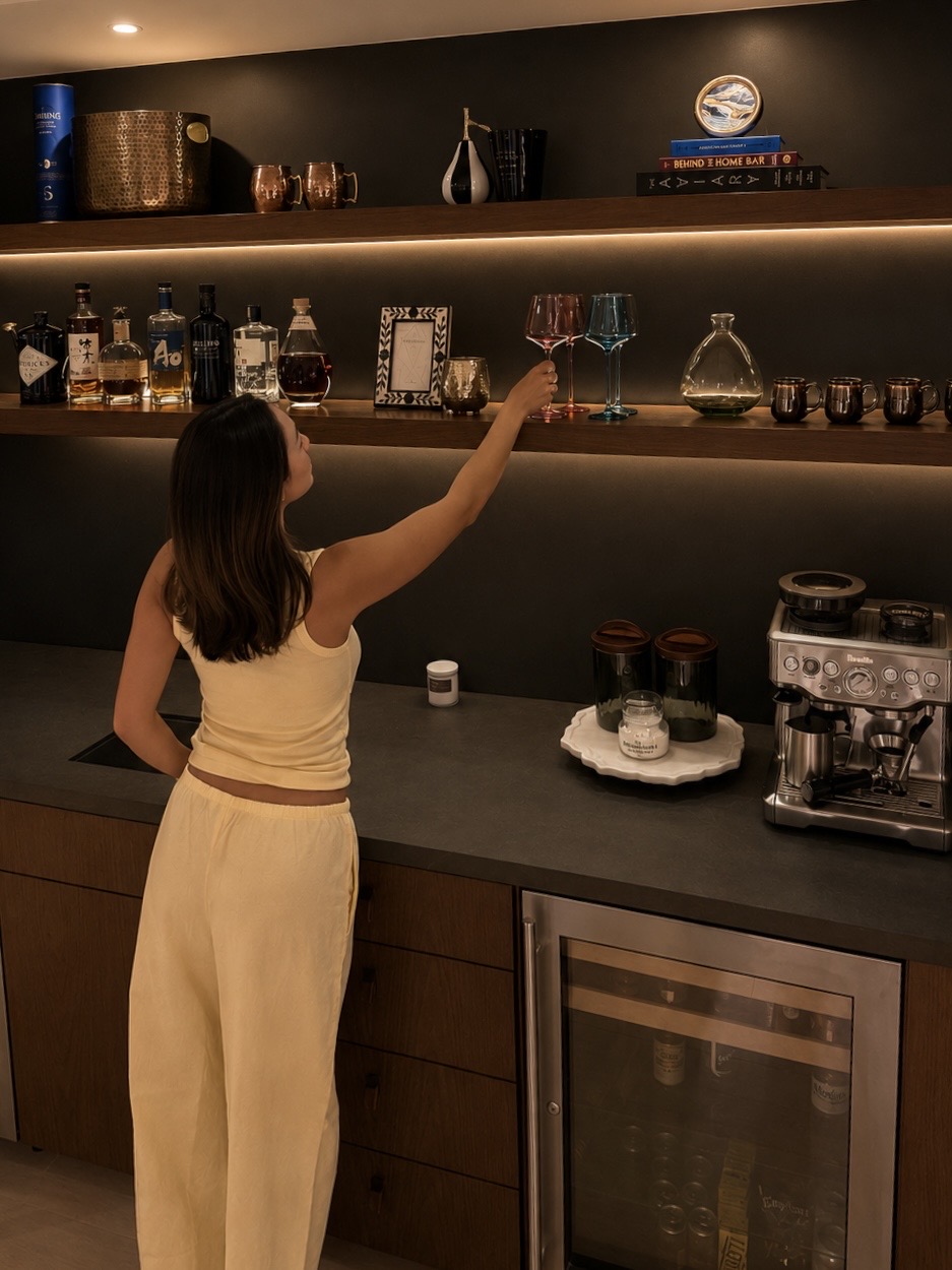 Styled home bar with warm under-shelf lighting, copper mugs, books and a brass bowl on dark floating shelves, liquor bottles and colorful glassware below, and a white tray of candles on the cleared counter