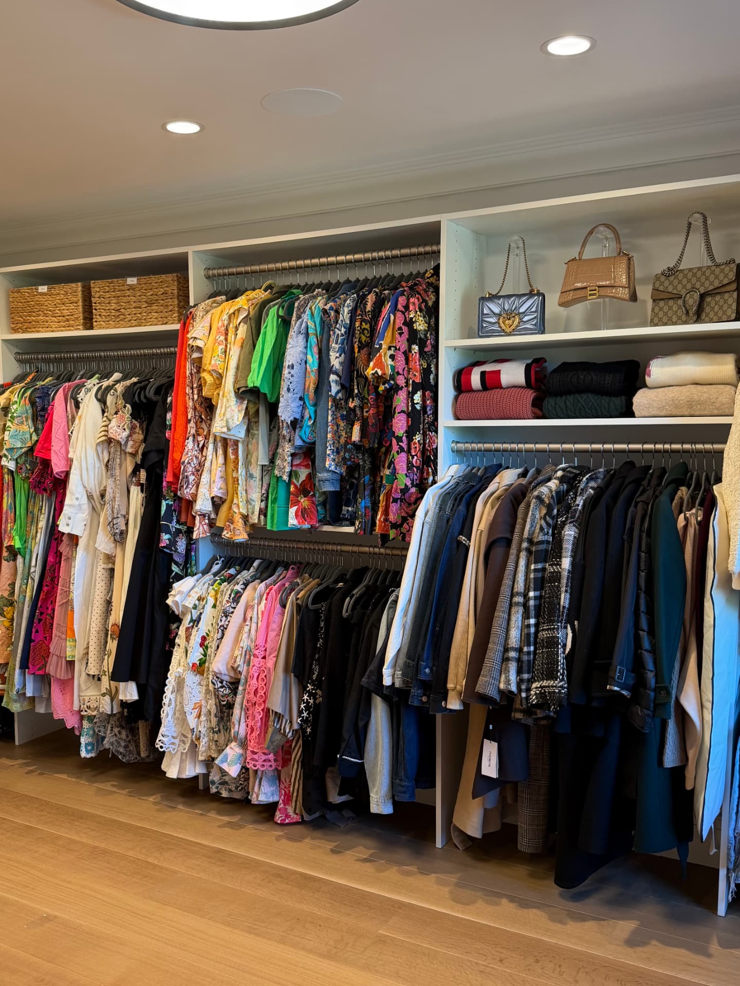 Wide view of the full wardrobe room with colorful dresses and tops hung by section, coats and jackets beside, handbags displayed on shelves, and woven baskets above