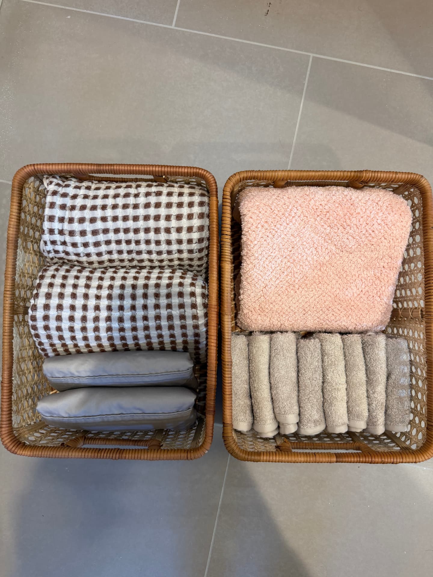 Two cane baskets on the floor holding rolled checkered textiles, gray pouches, a folded pink towel, and rolled washcloths