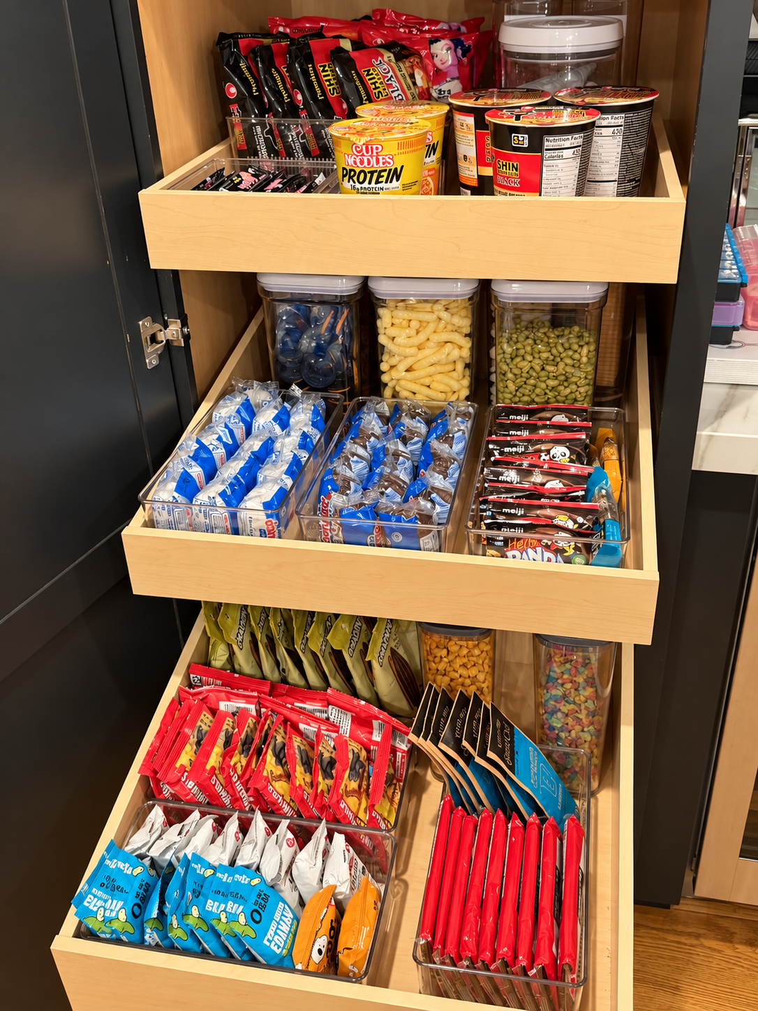 Three light wood pull-out pantry drawers with clear airtight containers and bins of snacks, cup noodles, and ramen packets