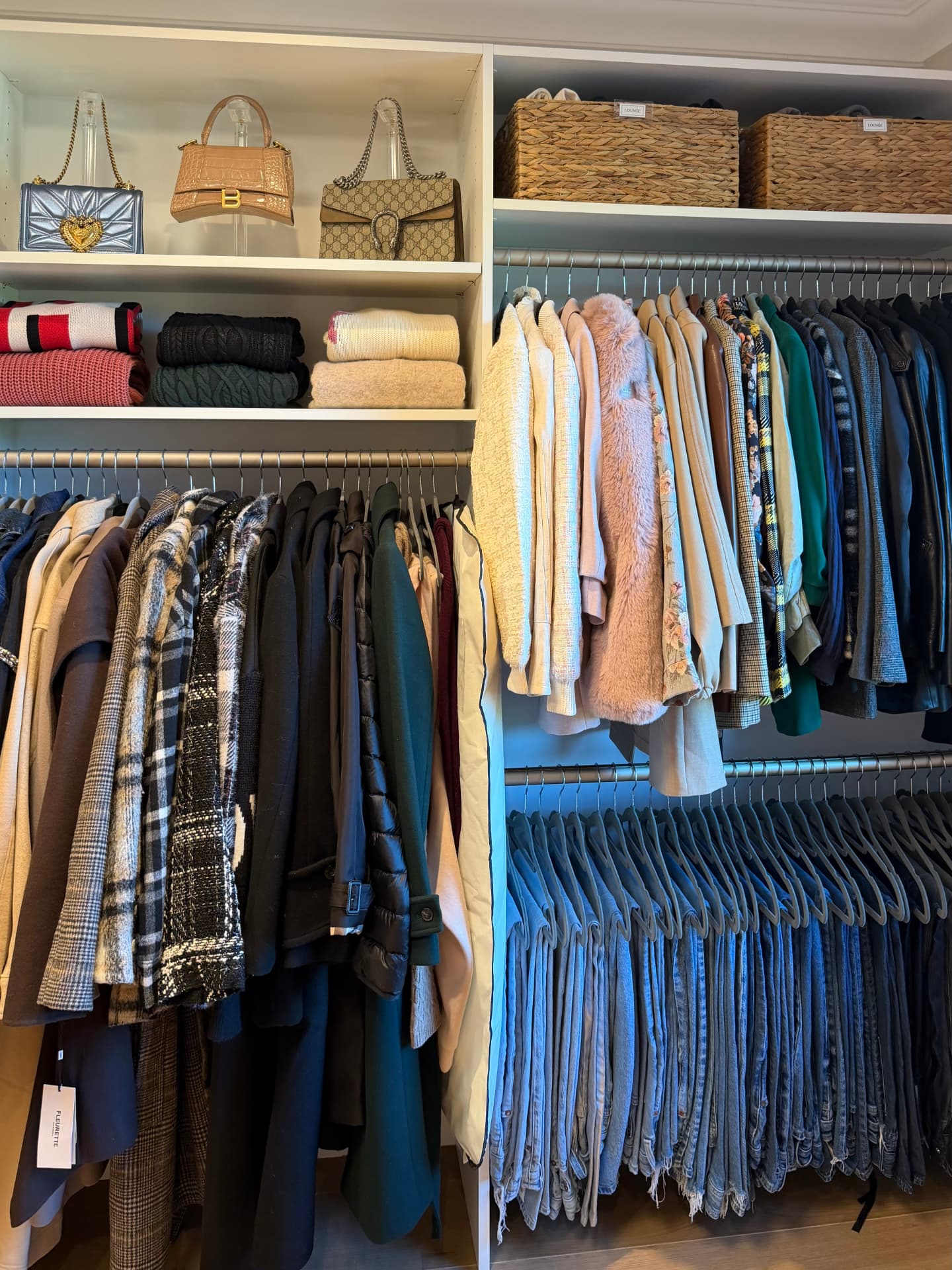 Organized wardrobe room with shelves of folded sweaters and handbags above a rail of coats and jackets, and color-sorted jeans hung below
