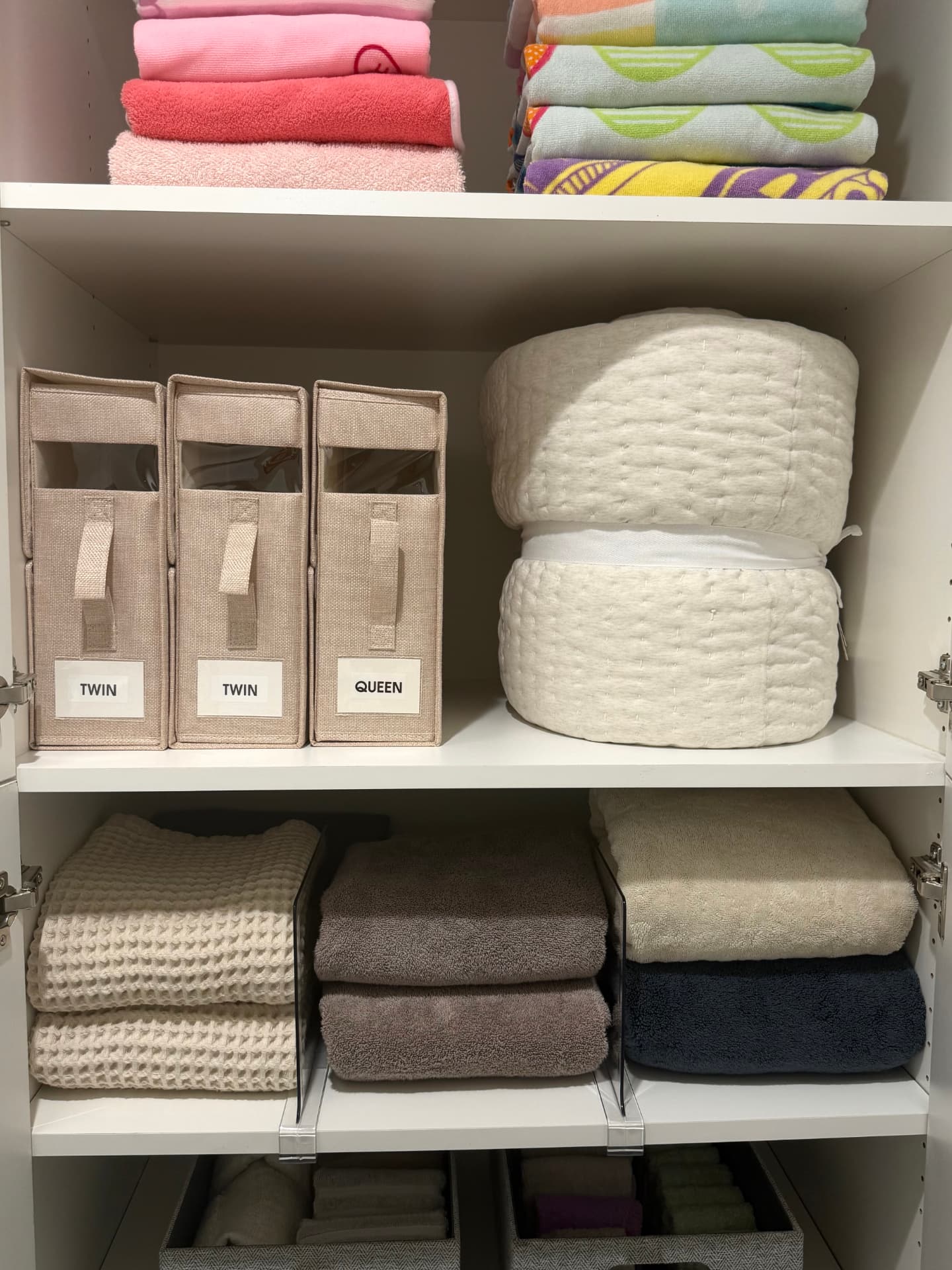 Organized linen closet with labeled twin and queen bedding bins, rolled comforters, and towels folded by color