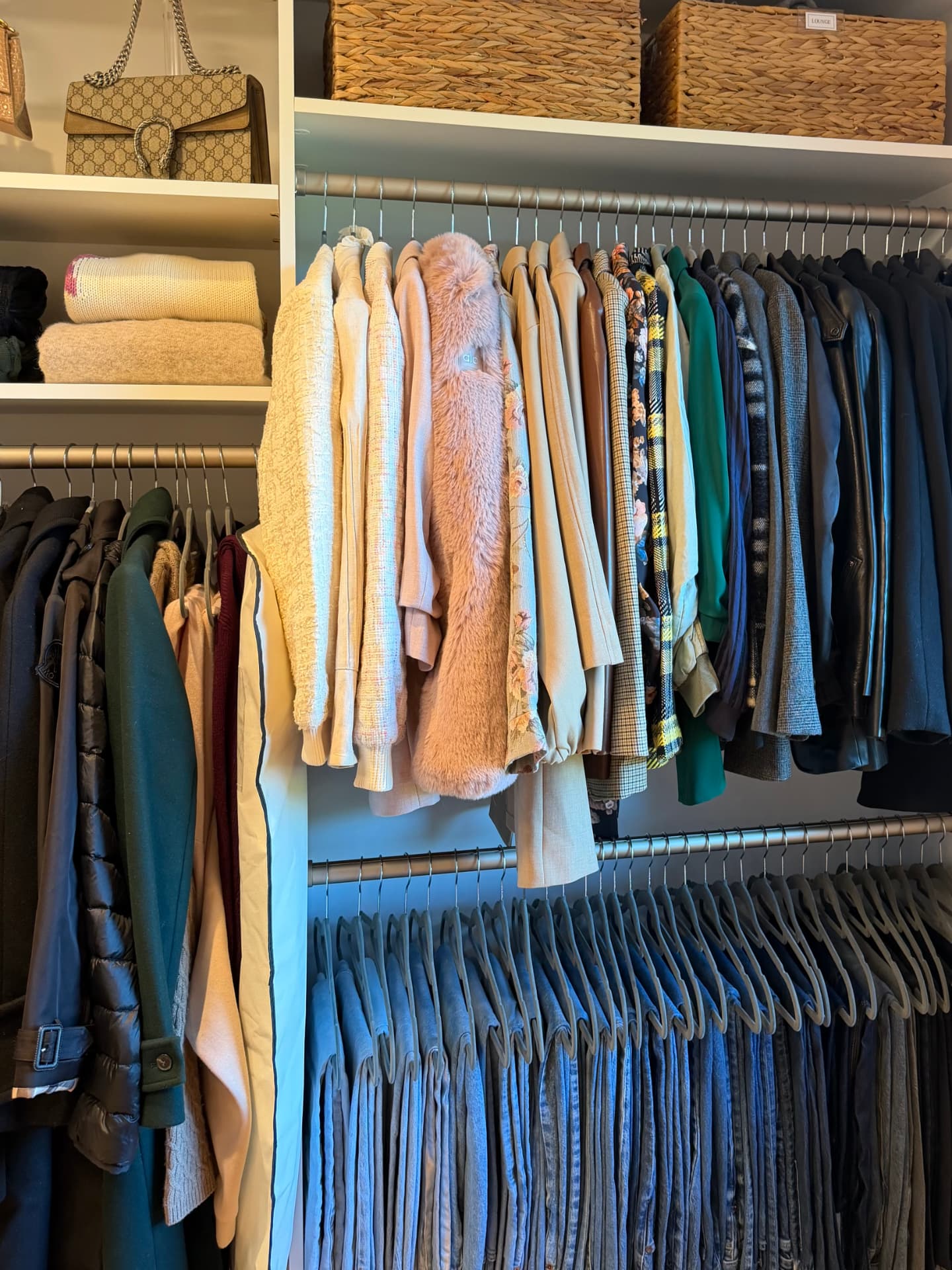Close view of a hanging rail with sweaters, jackets, and coats color-sorted, a handbag on the shelf above, and a rack of jeans below